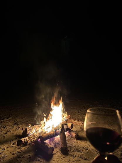 A bonfire on a beach with a glass of red wine in the foreground.