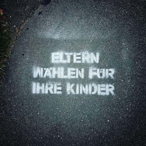 Mit weißer Farbe auf den Asphalt gesprühter Slogan „ELTERN WÄHLEN FÜR IHRE KINDER“.