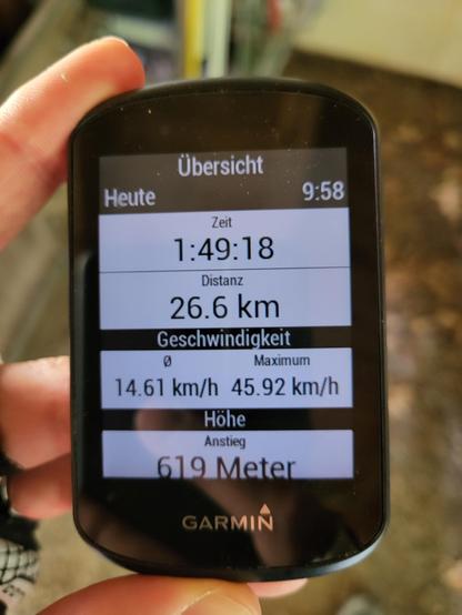 A person is shown in the image holding a cycling computer. The device is displaying various metrics such as the current time (9:58), duration (1:49:18), distance traveled (26.6 km), average speed (14.61 km/h), maximum speed (45.92 km/h), elevation gain (619 meters), and the brand "GARMIN". 