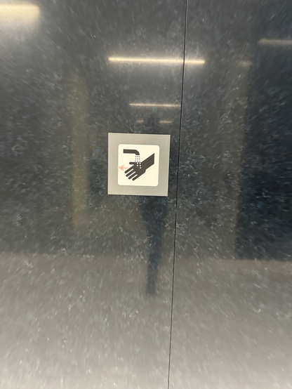 A white sign with a gray border against a dark gray granite wall. The sign shows a faucet sprinkling water drops onto a hand (and, it looks like, punching small holes in the hand). Someone has added an arrow using a pink marker pointing left, toward the sinks. Otherwise there’s no indication of where the sinks are.