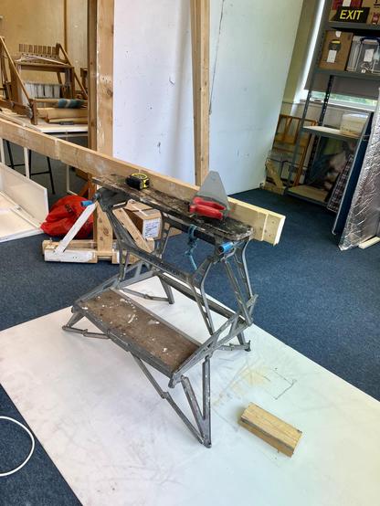 A workmate bench stands on a white sheet on the floor of the studio. A piece of timber is clamped in it and one end lies on the floor. A red handled saw lies on top of the bench. Behind, more wood is leaning up against the wall.