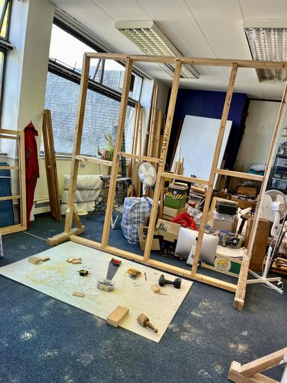 The timber frame of a partition wall stands in the studio. On a white plastic sheet in front of it is a pile of tools. Through the frame can be seen piles of stuff still covering the floor.