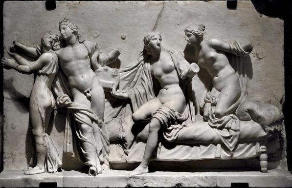 Marble relief of Apollon in a state of undress as he dances with a Muse in a long, flowing dress. On the bed behind him, two Muses have already undressed.