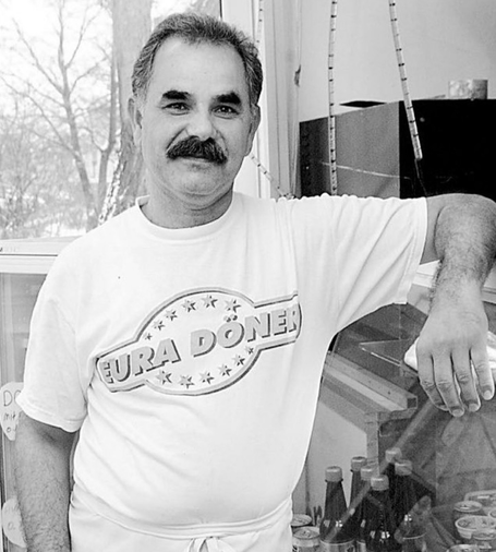 İsmail Yaşar lächelt in die Kamera. Vor allem seine Augen lächeln. Er trägt einen Schnauzbart. Ein weißes T-Shirt mit Aufschrift eines Döner-Ladens. Sein linker Arm lehnt an der Theke