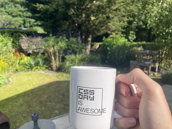 A hand holding a “CSS Day is awesome” memed white mug. In the background there’s a green sunny garden.