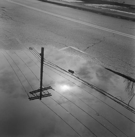 Square black and white photo of  utility wires, a telephone pole, and a cloudy sky reflected upside down in a puddle with some wet road surface visible in the upper half of the image.