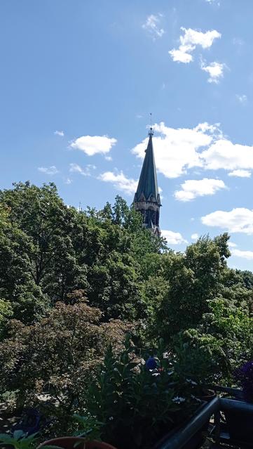Blick vom Balkon mit vielen Bäumen, ein spitzer Kirchturm, und Wolken, auf strahlend blauen Himmel, wie im Stil von René Magritte 