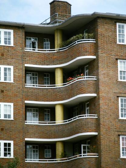 Low-rise brick apartment block in London, England, featuring small paned white windows and curvy balconies. The balconies seem to swing around a central pale gold pillar, at the top of which, on the roof, is a brick chimney.