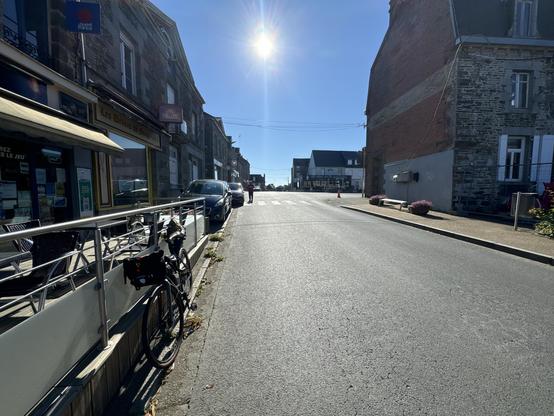 A view up a street. 
A bicycle rests against a metal barrier around some cafe tables. There are building on either side. An irregular shape of very blue sky with the sun bright - and some lens flare. 