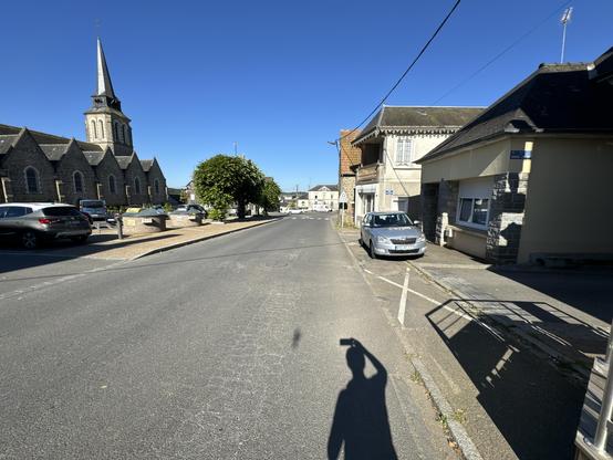 A view down the street. Shadow of the photographer in the foreground. Deep blue sky. 
A church to the left has a square tower with a small spire on top. There are five gables section visible on the side of the church. 
