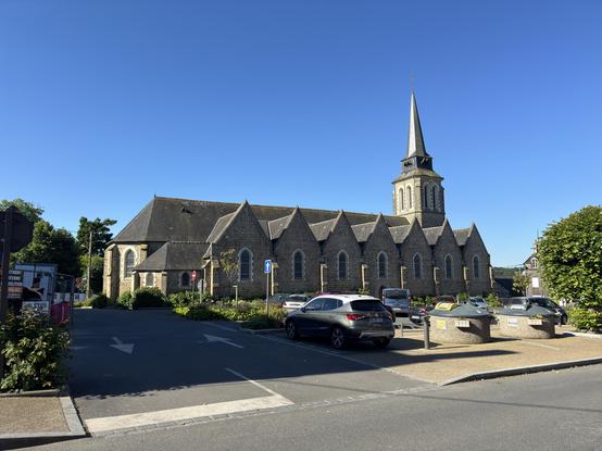A stone church across a strest with a square tower with a small spire on top to the right . There are seven gables section visible on the side of the church. 

There is a car parked in front of the church 
