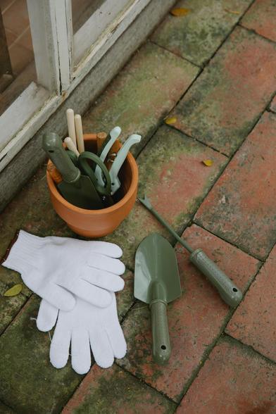 white gloves, trowel, and weed pick on brick patio next to clay pot of garden tools.
