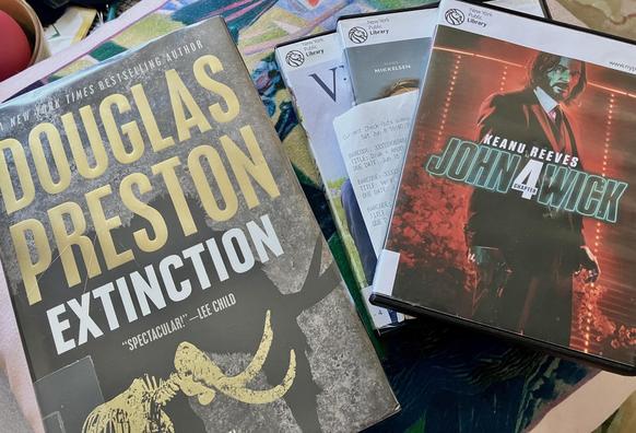 A book titled "Extinction" by Douglas Preston and a DVD case for "John Wick: Chapter 4" alongside other DVDs from the New York Public Library.