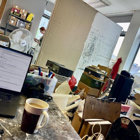 A mug of tea sits in the foreground on a table in front of a laptop. Behind can be seen a wall made up of two boards, one grey and the other messy white. There is still a pile of boxes in front of the wall.