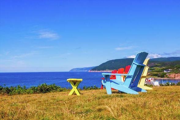Three vibrant blue yellow and red Adirondack lounge chairs and a small yellow side table invite you to relaxation overlooking the Pleasant Bay in Cape Breton, Nova Scotia, Canada in the summertime. 