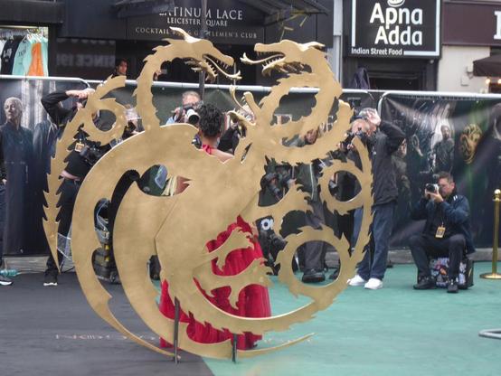 Targaryen dragon symbol in gold with, on the far side, actress in red dress posing for photographers.
