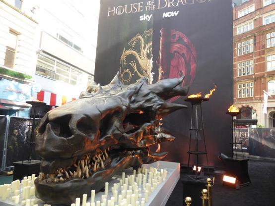Large, dark, dragon skull on low plinth, surrounded by unlit candles, with flame behind it, and House of the Dragon poster beyond that.