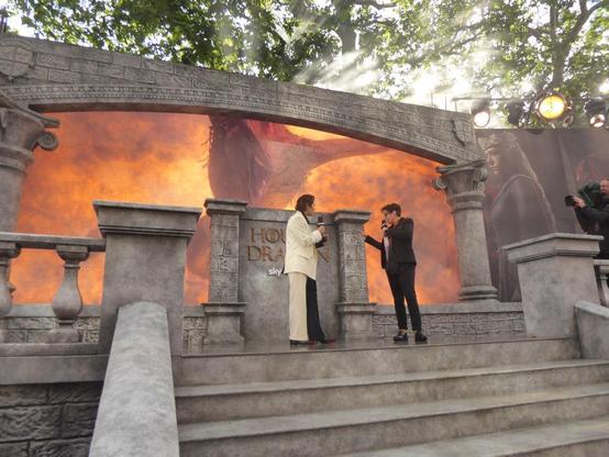 Emma D'Arcy in white and black suite being interviewed by Sue Perkins in black suit on stone-effect podium with video of a fiery dragon attack played on a screen behind them.