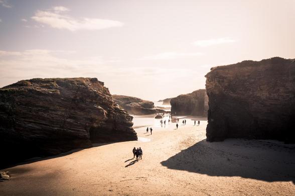 Fotografía desde un punto de vista ligeramente alto donde se ven figuras humanas caminando por la zona de arena y entre rocas de la Playa de las Catedrales en las primeras horas de la mañana.