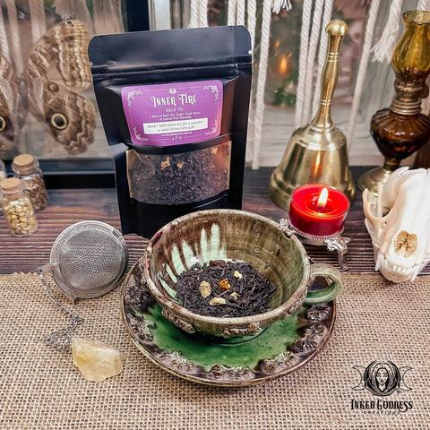 A pouch of loose leaf black tea stands behind a glazed earthenware teacup and saucer with a mesh tea infuser ball. The cup contains a portion of the herbal mixture. The pouch has a clear window on the front with a label that features the name of the tea, "Inner Fire," ingredients, and preparation instructions. This is an exclusive product from Inked Goddess Creations.
