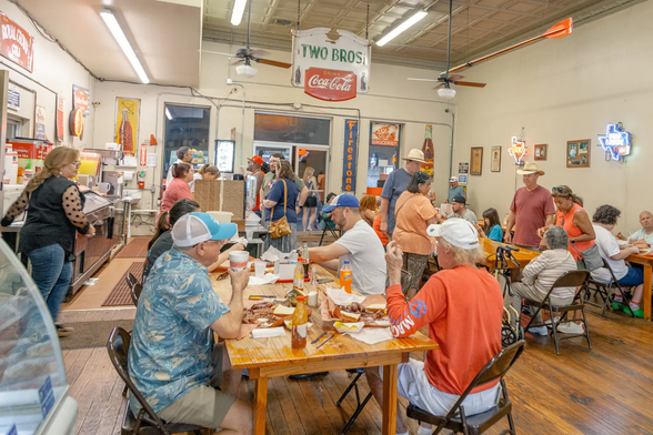 The dining room at Smitty’s Market, where generations of Lockhart families have gathered. Credit...Katherine Squier for The New York Times

Texas Diners dressed casually, many wearing baseball caps or cowboy hats sit on metal folding chairs at simple wooden tables enjoying delicious Texas barbecue. On the walls are throw-back signs from yesteryear and neon Texas beer lights. Beyond the diners in the background, customers are in line ordering their food at the counter. 
