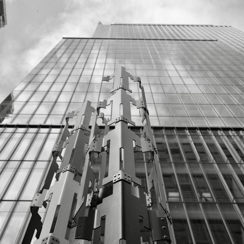 A black and white film photo. A permanent installation made of painted metal called Human Structures by Jonathan Borofsky stands in front of an office building in San Francisco, California. Friday, June 7, 2024.