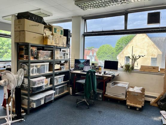 A pair of shelves are now up against the latest wall in the studio and are packed with boxes. A bench is next to them looking out of the window. It has a pair of screens on it and a laptop. A large printer sits on the floor under one edge. It’s bright outside and some trees and houses can be seen.