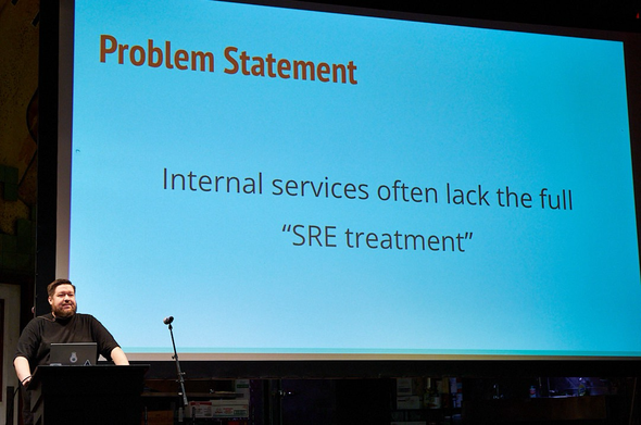 Speaker in front of screen with problem statement: internal services often lack the full “SRE treatment”