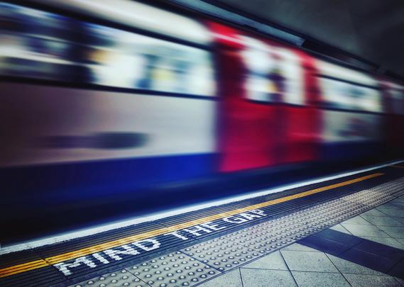 Ein typischer weiß-rot-blauer Zug der Londoner U-Bahn verlässt die unterirdische Haltestelle. Im Vordergrund ist die Kantenmarkierung des Bahnsteigs „MIND THE GAP“ scharf zu sehen, der ausfahrende Zug ist durch Bewegungsunschärfe nicht im Detail zu erkennen. 