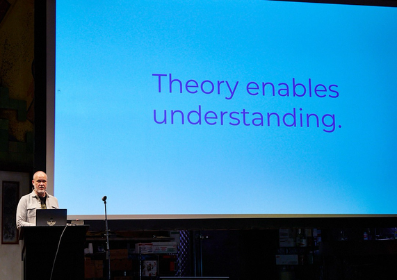 Speaker at a lectern in front of a screen that says “Theory enables understanding.”