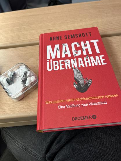 Ein rotes Buch mit dem Titel "Machtübernahme" von Arne Semsrott auf einer Holzoberfläche neben einem Plastikbehälter mit Ohrhörern im Inneren.
