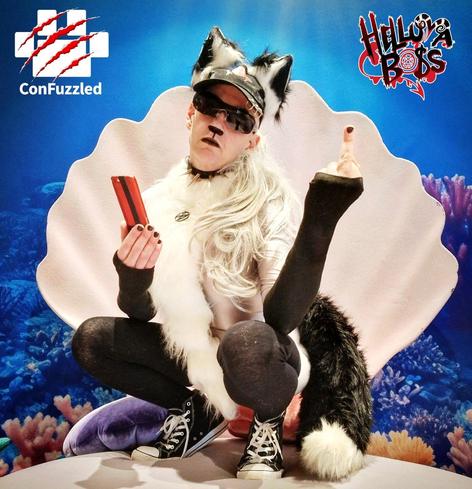 Cosplay of Loona from Helluva Boss, a grey and white wolf hellhound, squatting in a giant clamshell at an undersea photoshoot, holding a red mobile phone and making a rude gesture with her middle finger.
