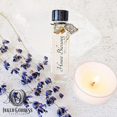 A small square vial of House Blessing Spell Oil is propped up against a large clear quartz point. It has a silver door charm tied to the neck of the vial. There sprigs of Lavender and a white tealight candle for scale. This is an exclusive product from Inked Goddess Creations.