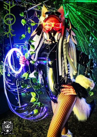 Cybergoth fox cosplay holding a glowing fibre optic whip standing in front of green laser beams and green hanging vines. Red glowing visor and black and white falls. White furry tail and armbands, black pvc bodysuit.