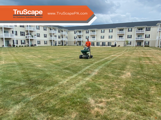 "Two photos showing the transformation of a commercial property lawn. The first displays a lush green lawn with striped patterns and residential buildings in the background. The second shows a TruScape worker using a fertilizer spreader on the same property, highlighting the improved grass condition. Both images feature a translucent orange overlay at the top with the TruScape logo, website www.TruScapePA.com, and the tagline 'An Outdoor Solution Provider.'"