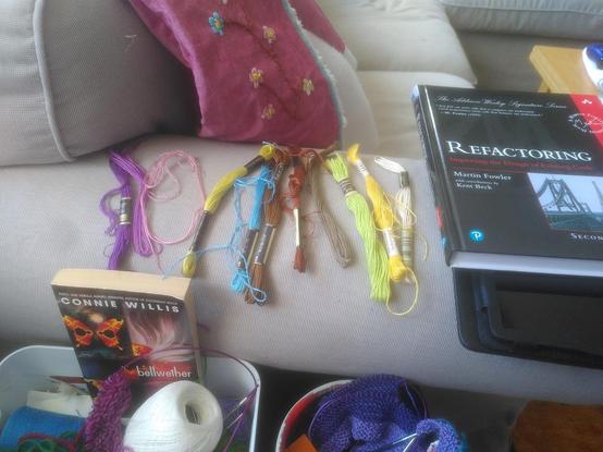 Photograph of the arm of a light grey sofa with embroidery floss in ten colors laid out on it, with a half-finished red/pink vest hanging over the back of the sofa. Besides the floss, a tablet PC and Martin Fowlers and Kent Becks classic book on Refactoring existing code reside on the sofa arm.
Below this is a box of knitting and sewing bric a brac and the novell Bellwether by Connie Willis. Left of the box is a knitting bowl with thin carbone needles and purple purl knitting.