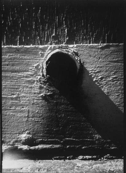 A black and white pinhole picture of a drain hole in a wall with a rusty stain underneath