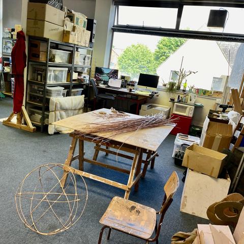 A trestle table stands in the middle of the studio space. On it lies a bundle of willow withies. In front of the table on the floor is a roughly spherical shape made out of curved and taped pieces of willow.