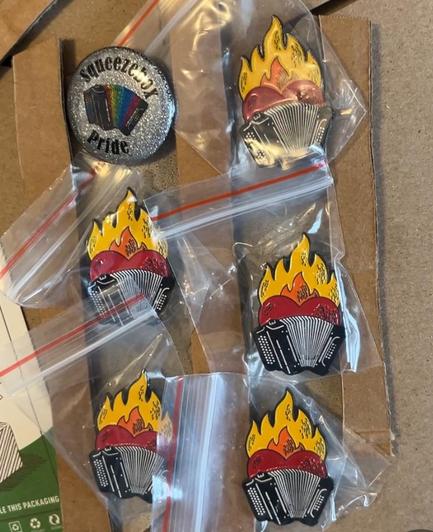 Flaming-heart Accordion enamel pins individually wrapped in tiny ziplock bags. Prepared for shipping taped with recycled cardboard backing, next to a glittery rainbow accordion badge with the text “squeezebox pride“. 