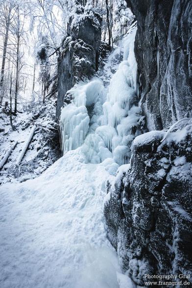 A stunning photograph of a frozen waterfall captured on a rocky cliff is showcased in this image. The frozen waterfall appears majestic against the rocky backdrop, creating a beautiful contrast between the icy white cascades and the dark, rugged rocks. The scene exudes a sense of tranquility and serenity, with the frozen water suspended in time. The image captures the essence of winter, with snow-covered trees in the background adding to the wintry atmosphere. The overall color scheme is domina…