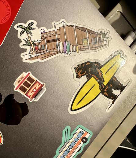 Stickers on a computer. Main focus is a sticker with a picture of the Taco Bell in Pacifica California. The Taco Bell is very popular and right on the beach.