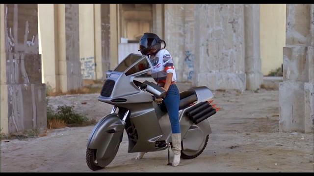 A person wearing a helmet stands next to a futuristic motorcycle in an urban, industrial setting.