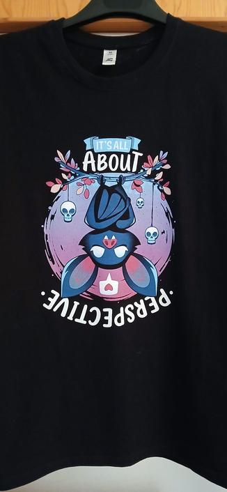Camiseta negra colgada de una percha. Dibujo de lindos colores de un  murciélago bocabajo con las alas cruzadas. Cuelga de una rama con hojas de tonos rosas y 3 calaveras penden de un hilo. 'It's all about perspective'