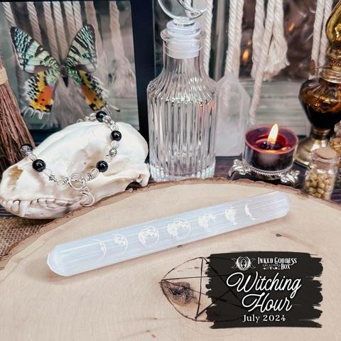 A selenite wand sits on a raw wood slice. The wand is cylindrical with rounded ends. It has seven moon phases etched along most of its length on one side This is one of the items in the exclusive July 2024 "Witching Hour" Inked Goddess Creations Box.