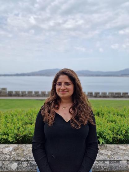 Dr Milia Helena Hasbani smiling, wearing glasses a black jumper, stood in front of a grey stone wall, with green bushes behind her and a body of water in the distance.