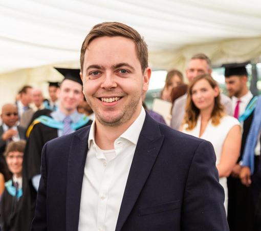 Dr Benjamin Metcalfe wearing a white shirt and dark blue suit jacket, smiling. In the background are people wearing formal wear and graduation gowns.