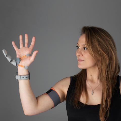 Dani Clode, wearing a black top. Her right hand is raised on it there is a grey and orange extra robotic digit (from her ‘Third Thumb’ project) and she has a grey strap around her upper arm.