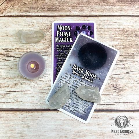 A pair of cards are arranged on a distressed wood surface. Each card in the "Moon Phase Magick Expansion Pack for the The Eclectic Witch Card Deck" product features information on a different moon topic; namely, Moon Phase Magick and Dark Moon Magick. This is an exclusive product from Inked Goddess Creations.