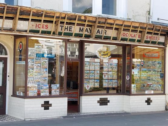 An estate agents shop with various properties displayed in the windows. Above these is a hand-painted fascia sign for a former occupant, a confectioner called The Mayfair. A wooden structure has been erected over this which at one time allowed a larger sign to be mounted, but only its supports remain, making the older sign visible beneath.