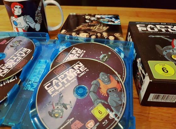 Das Foto zeigt die geöffnete Box der Captain-Future-BluRays der "Uncuts" (also der ungeschnittenen Fassung), den Box-Schuber, eine Captain-Tasse sowie im Hintergrund die Firefly-Box